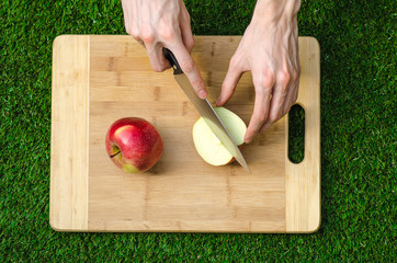 Vegetarians and cooking on the nature of the theme: human hand holding a knife and a red apple on the background of a cutting board and green grass top view