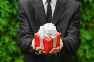 Wall Mural - Gift and business theme: a man in a black suit holding a gift in a red box with a white ribbon on a background of green grass