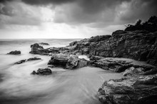 Black And White Image Of Wave Hitting The Rock. Dark And Dramatic Clouds. Soft Focus Due To Long Exposure