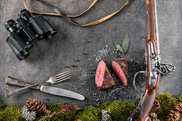 deer or venison steak with antique long gun, cutlery, binocular and ingredients like sea salt, herbs