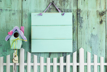 Blank mint green sign hanging over white picket fence and colorful birdhouse