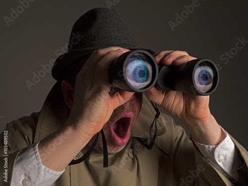 Fotografía Photograph of a man in trench coat and hat looking through binoculars with huge, cartoonish eyes seen in the lenses