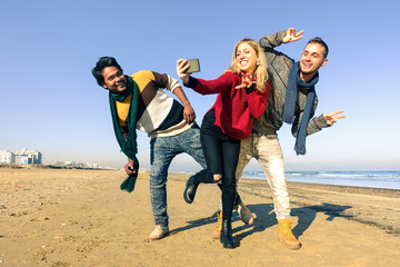 Wall Mural - Multiracial group of friends having fun dancing on the beach while taking selfie - Young funny people joyful on a sunny spring day waiting for summer time - Concept of happiness and teenage travel 