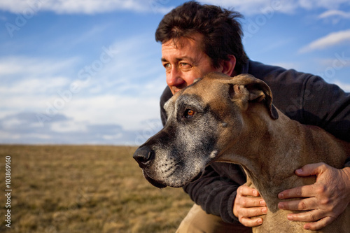 Fotografia, Obraz Rugged man holding his great Dane in a windswept field with dramatic sky