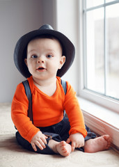 Portrait of cute adorable stylish Caucasian baby boy with black eyes in hat, orange shirt onesie, jeans with suspenders barefoot sitting on windowsill looking in camera, natural window light