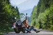 Handsome biker with beard and long hair sitting next to a traveler motorcycle on an open road. Guy is wearing leather jacket and blue jeans. Sunny summer day in the mountains.