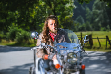 Portrait of biker with long hair and beard in a leather jacket and sunglasses sitting on his bike beside the road in the forest. Looking into the camera. Tilt shift lens blur effect