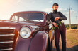 © endrews21 - brutal bearded man with a mustache in a shirt, pants with suspenders stands leaning on the hood of a retro car holding a white rose and looking at the sunset
