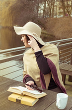 Pretty French Brunette Woman In Hat And Poncho Reading Vintage Book And Drinking Coffee Waiting Spring Near Lake. Early Spring.