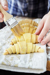 Chef smearing raw croissants with brush in yolk, preparation process. Dough for croissant sweet traditional dessert.