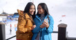 © Dash - Two cute smiling young adult women in long black hair brown and blue jackets looking at a cell phone while standing in front of a ski slope.