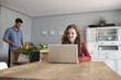 © Westend61 - Woman at home using laptop with man in background cutting vegetables