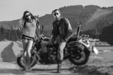 Couple standing by a motorbike. Handsome man wearing leather jacket, gloves and boots and young beautiful sexy woman wearing shorts. Summer day. Tilt lens blur effect. Black and white