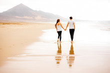 Young And Lovely Couple In White T-shirts And Black Pants Walking Beautiful Sandy Beach With Mountains On Background On Fuerteventura Island In Spain. General Plan With Copy Space