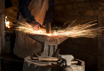 The blacksmith manually forging the molten metal on the anvil in smithy with spark fireworks