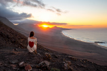 Young woman enjoying beautiful sunset sitting on the mountain with great view on Cofete coastline on Fuerteventura island