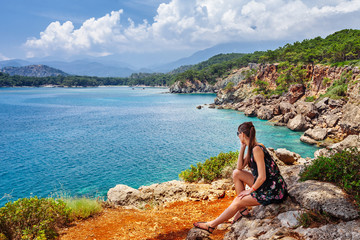 Wall Mural - Portrait of beautiful tanned woman sitting at the sea coast. Hot summer day and bright sunny light. Panoramic view on sea shore near Kemer, Antalya, Turkey.