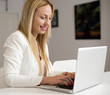 © Freestocker - Young woman with blond hair wearing white jacket smiles while working with her laptop in the office. Business