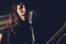 Pretty Woman Singing With Guitar. Attractive Female Playing An Electric Guitar And Singing Into A Vintage Microphone. Set In Dark Room With Spot Lighting And Edited With Vintage Effects.