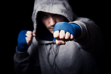 A boxer in hood jacket and bandages punches. Close up portrait in the dark.