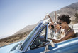© Mint Images - Three young people in a pale blue convertible car, driving on the open road across a flat dry plain,