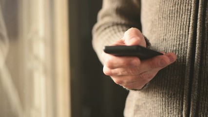 Wall Mural - Man using mobile smartphone for texting messages, adult male person standing by the window of dark living room sending SMS with smart phone app, selective focus with shallow depth of field.