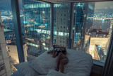 Young beautiful woman lying in the bed,making selfie or reading messages or chatting at night.Luxurious apartment with french windows,breathtaking view over harbour,skyscraper.Addiction to internet
