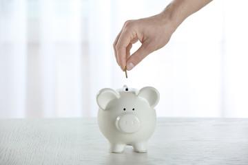 Poster - Male hand putting coin into white piggy bank on blurred curtains background