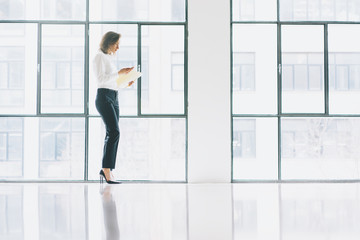 Wall Mural - Photo business woman wearing modern suit, looking  mobile phone and holding papers in hands. Open space loft office. Panoramic windows background. Horizontal mockup.