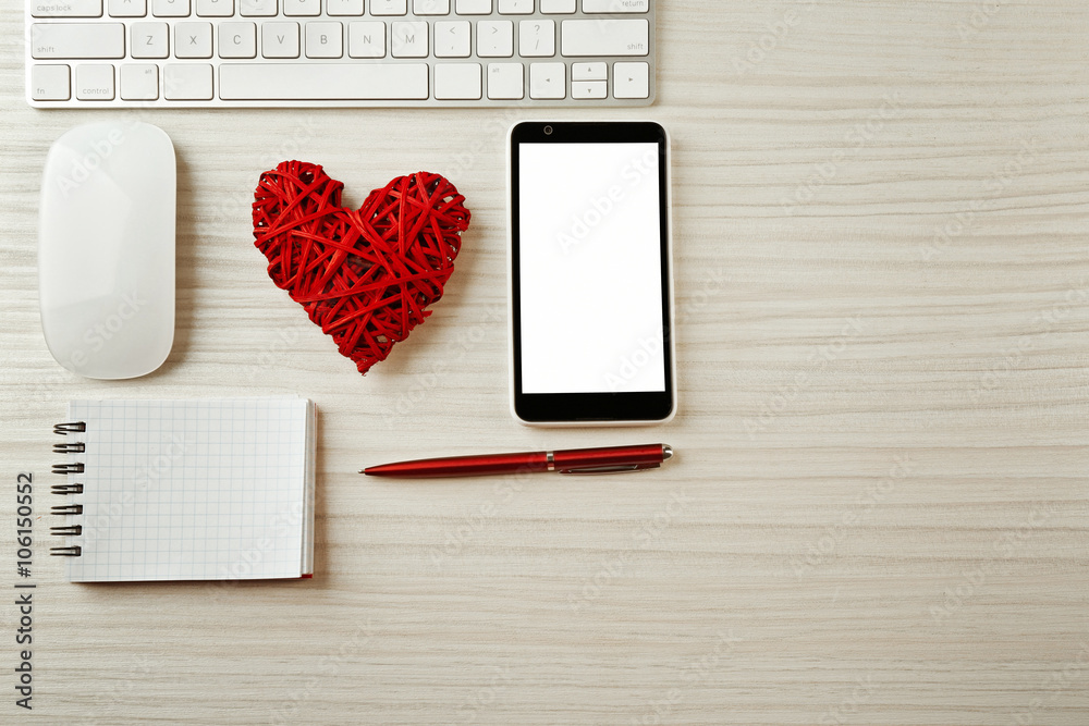 Computer peripherals with wicker red heart, notebook and mobile phone on light wooden table
