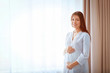 © Africa Studio - Pregnant woman standing near window with curtains in the room