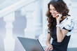 © GTeam - Cute business woman brunette with long curly hair,dressed in a black dress with white sleeves,sitting on the steps near the white railing,works with laptop and talking on mobile phone outdoors