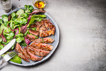 Sliced grilled beef steak with green leaves salad on rustic plate with cutlery. Medium rare barbecue steak and healthy salad on gray stone background, top view, place for text