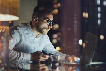 Wall Mural - Bearded young businessman working on modern loft office at night. Man using contemporary notebook texting message, holding smartphone, bokeh background. Horizontal, film effect. 