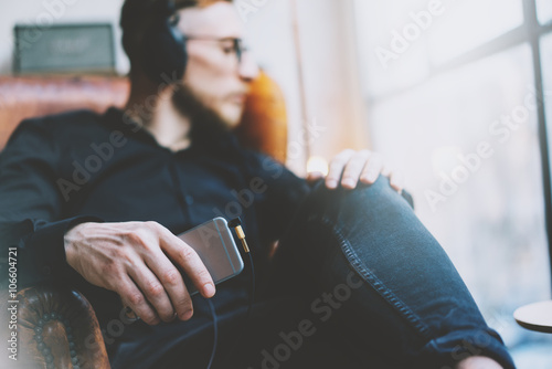 Foto Hübsche bärtige Mannkopfhörer des Fotos, die Musik modernes Dachbodenstudio hören Mann, der im Weinlesestuhl schaut Fenster sitzt, Smartphone hält und sich entspannt Unscharfer Hintergrund horizontal, Filmeffekt