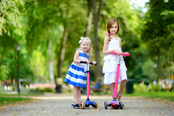 Wall Mural - Two adorable little sisters wearing beautiful dresses riding their scooters