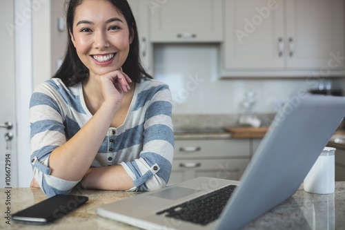 Portrait of young woman sitting with laptop and mobile phone in kitchen at home Poster