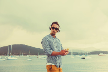 Wall Mural - stylish young man in a shirt and shorts and sneakers listening to music in headphones on a smartphone and is sitting on pier looking at sea on yachts boats and the sunset,music lounge ,relaxing 