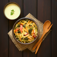 Canvas Print - Baked tricolor fusilli pasta and vegetable (broccoli, tomato) casserole in rustic bowls, wooden spoon, fork and cream sauce on the side, photographed overhead on dark wood with natural light