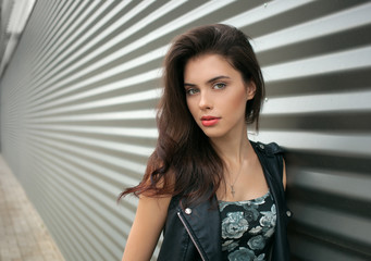 Wall Mural - Closeup portrait of young beautiful brunette woman in black leather jacket posing outdoors against urban style background of metal strips