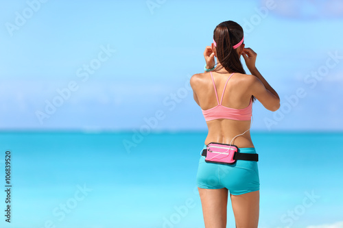 Fotografía Chica corredor de fitness está preparando para la ejecución en playa escuchando música con auriculares y soporte de teléfono inteligente correa de cintura riñonera como ropa deportiva que llevan portátil rastreador de actividad de tecnología