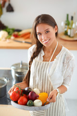 Wall Mural - Smiling young woman holding vegetables standing in kitchen