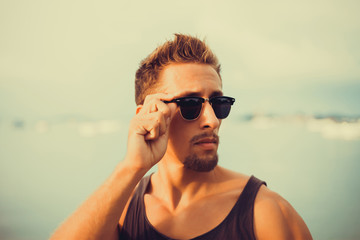 Wall Mural - young guy with a beard in a t-shirt and sunglasses posing on the pier,Rugged and manly. Confident young bearded man looking at camera and adjusting eyewear while standing outdoors