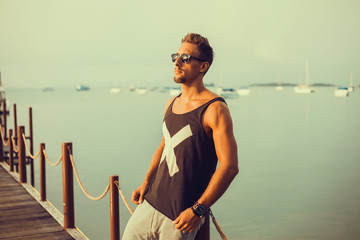 Wall Mural - young guy with a beard in a t-shirt and sunglasses posing on the pier,Rugged and manly. Confident young bearded man looking at camera and adjusting eyewear while standing outdoors