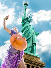 A Smiling And Fashionable Woman With A Orange Wide-brimmed Hat Takes A Selfie. Statue Of Liberty And Blue Sky In The Background. Liberty Island, New York City, United States.