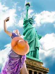 A smiling and fashionable woman with a orange wide-brimmed hat takes a selfie. Statue of Liberty and blue sky in the background. Liberty Island, New York City, United States.