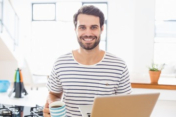 Poster - Portrait of happy man holding coffee cup and laptop in kitchen