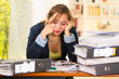 © Fotos 593 - Business woman sitting by desk, paper files spread out, leaning head on hands looking overwhelmed and tired