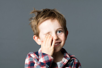 closeup portrait of a smiling young red-hair child and blue eyes hiding one eye for fun eyesight or joyous hide-and-seek, grey background studio