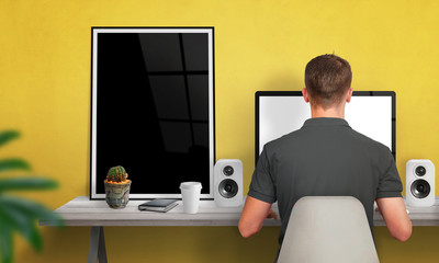 Poster - Man work on computer. Poster frame beside. Coffee, cactus, speakers on table. Free space on wall for text. Yellow wall in background.